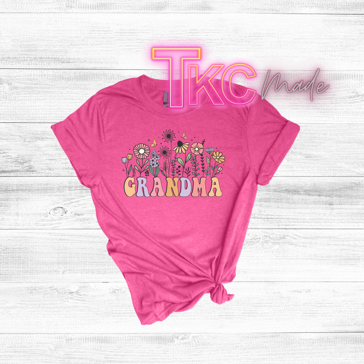 Pink t-shirt with 'TKC Grandma' text and floral designs on a light wooden surface.
