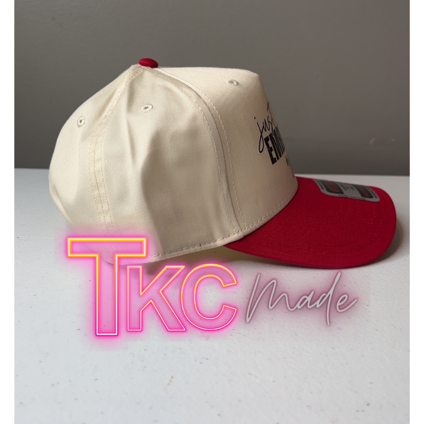 Two baseball caps, one beige and one red, on a gray surface with 'TKC made' text.