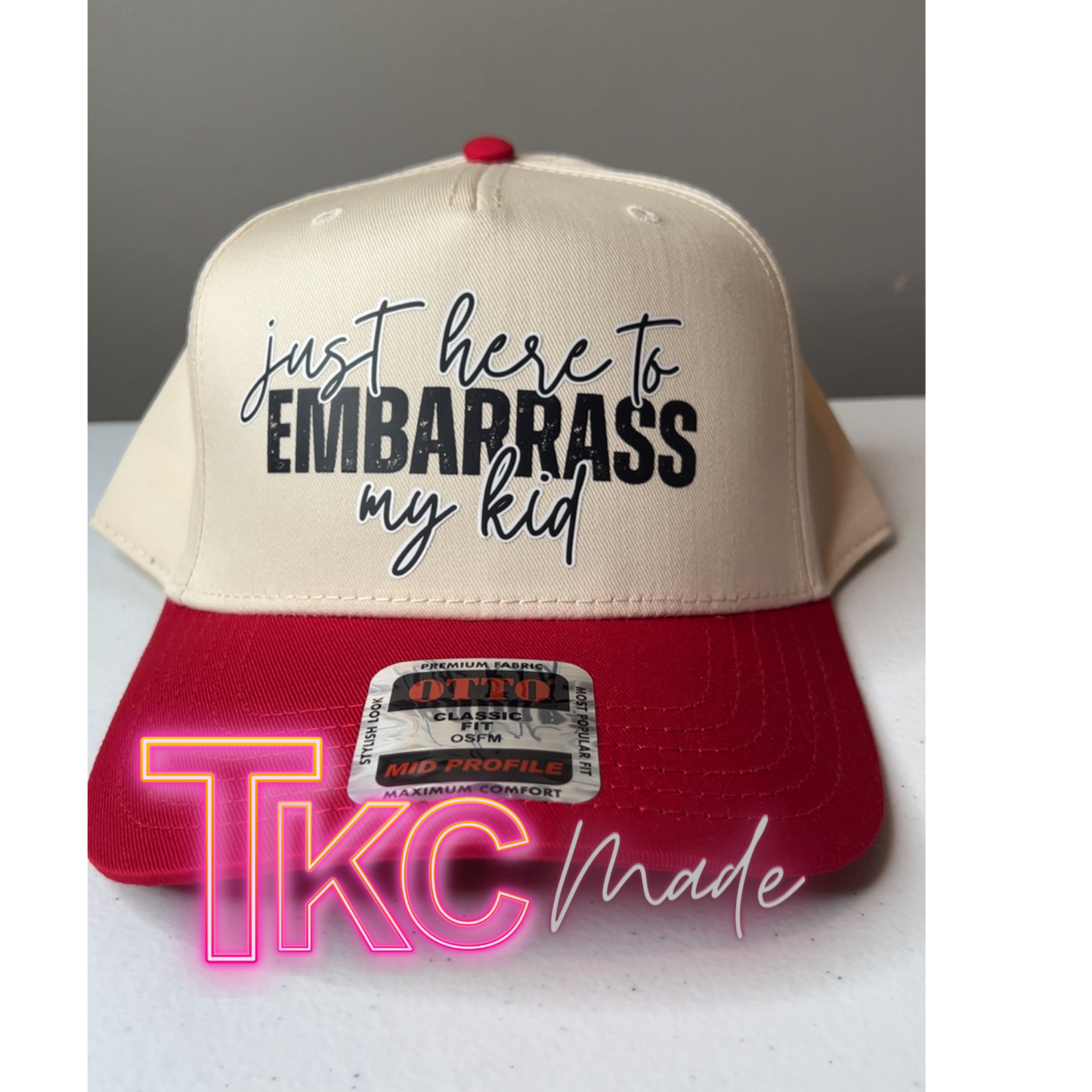 Beige and red cap with text 'just here to embarrass my kid' on a gray background