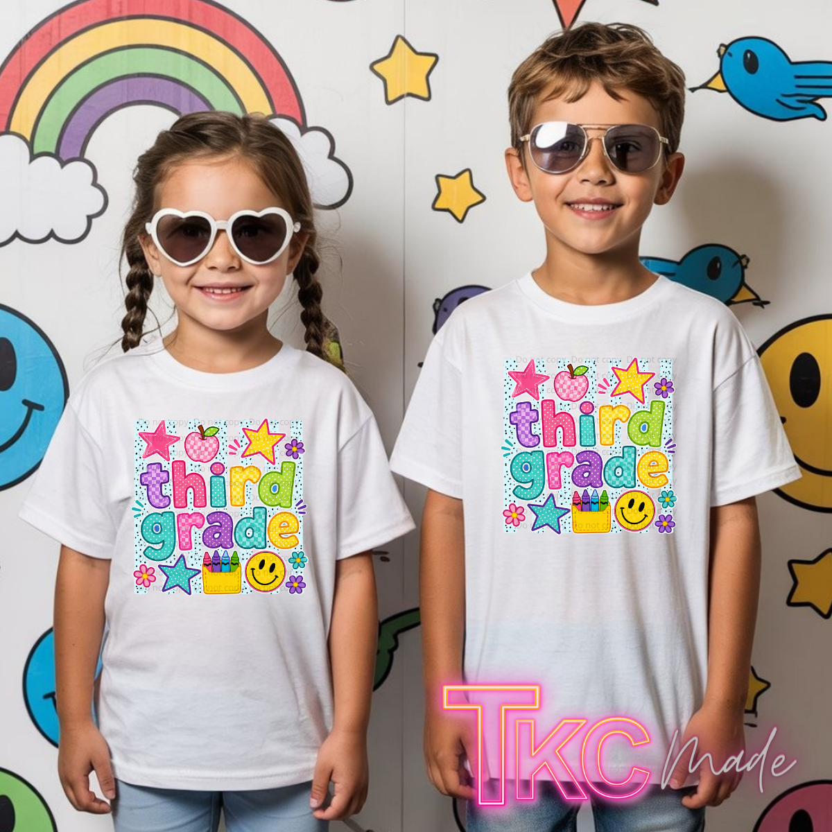 Two children standing side by side wearing matching white graphic t-shirts with a colorful 'third grade' text design, set against a playful wall with cartoon drawings.
