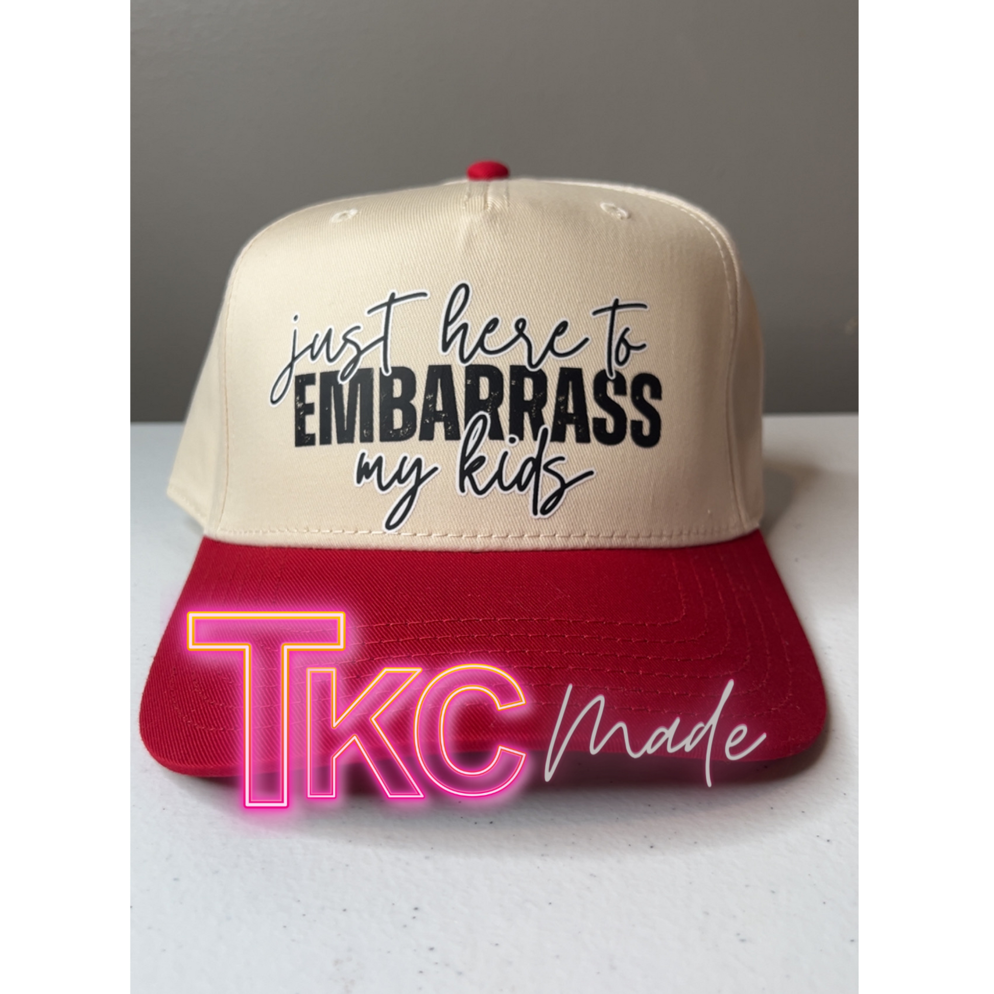 Beige and red cap with text 'just here to embarrass my kids' and 'TKC Made' on a gray background