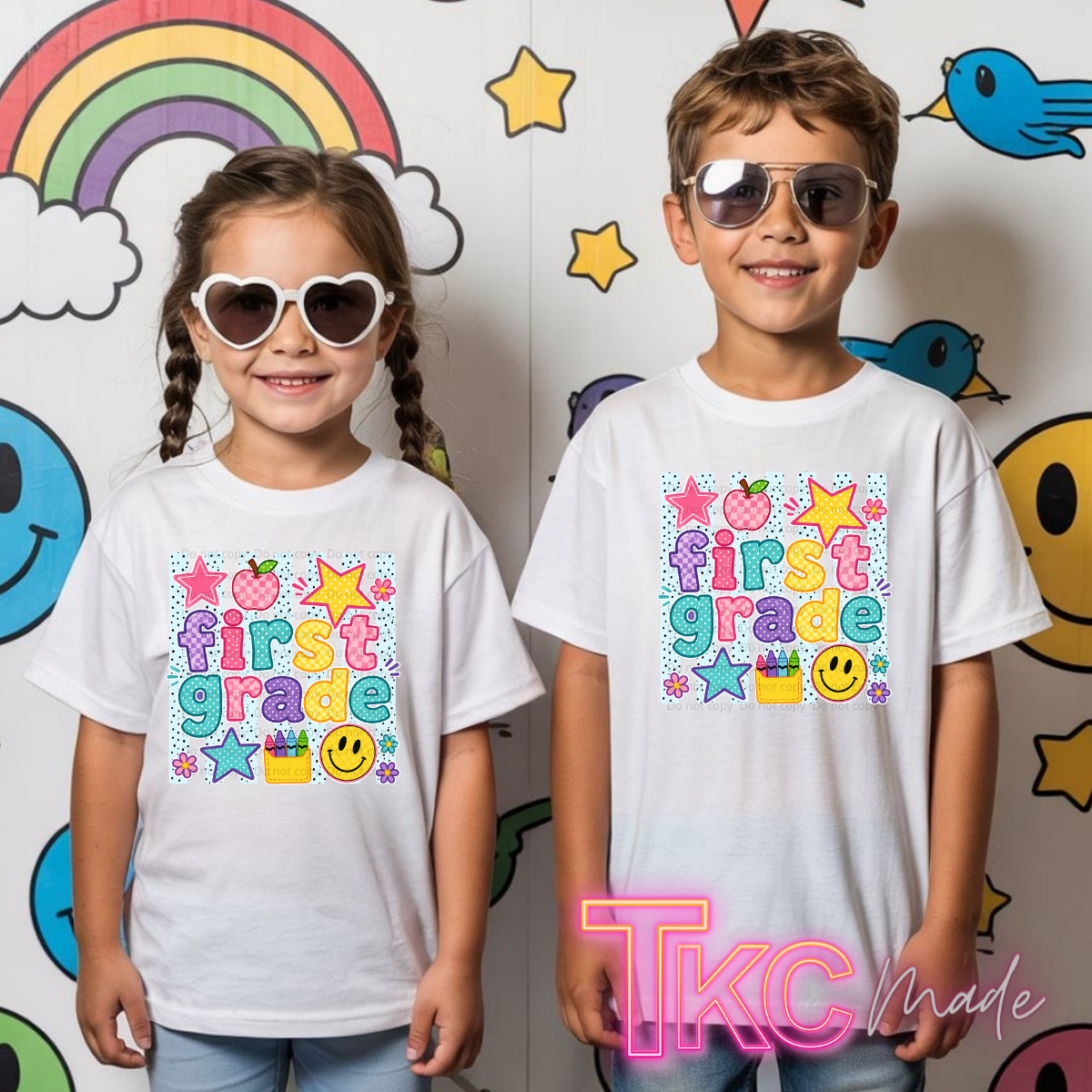 Two children standing side by side wearing matching white graphic T-shirts with a colorful 'first grade' design, posed in front of a playful wall decorated with cartoon smiley faces, stars, a rainbow, and birds.