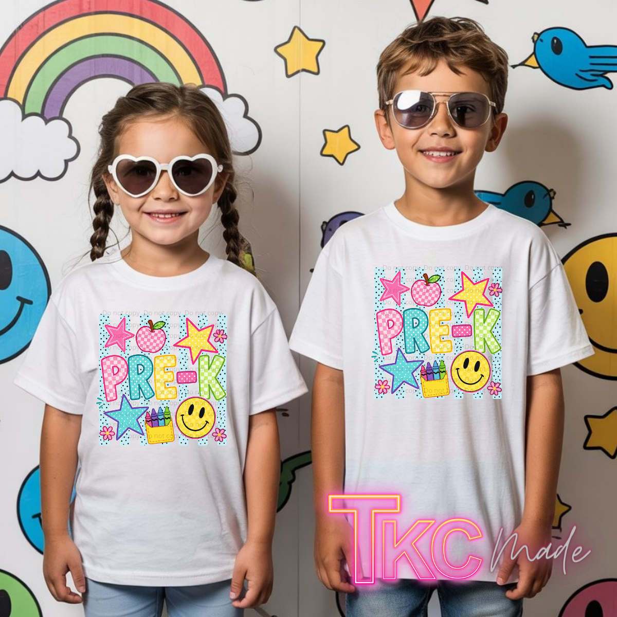 Two children, a girl and a boy, wearing matching white graphic t-shirts with a colorful 'PREK' design. The background is a playful wall decorated with cartoon smiley faces, stars, a rainbow, and birds.