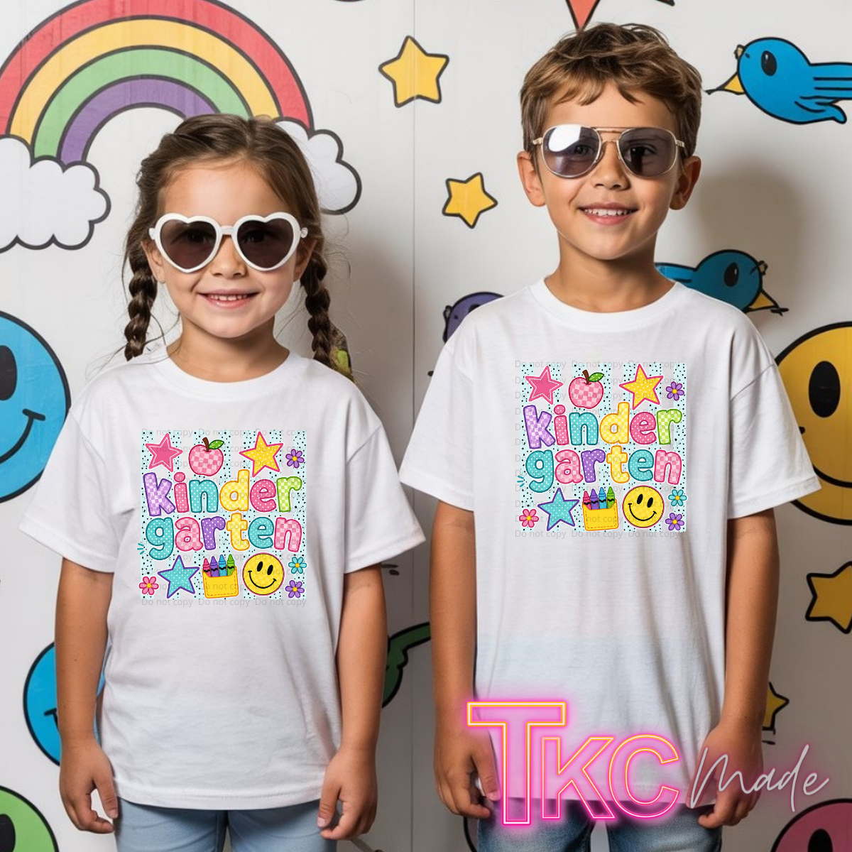 Two children standing side by side wearing matching white graphic T-shirts with a colorful 'Kindergarten' design, posed in front of a playful wall decorated with cartoon smiley faces, stars, a rainbow, and birds.
