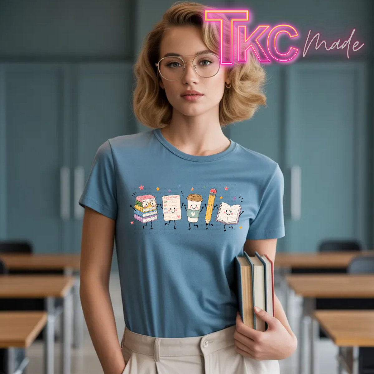 A woman wearing a light blue graphic t-shirt with a cute cartoon school-themed print, standing in a classroom setting holding books. A pink 'TKC made' watermark is visible in the top right corner.