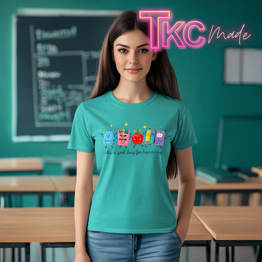 A woman stands in a classroom wearing a teal graphic T-shirt with colorful cartoon characters and the text 'It's a good day for learning.' A large pink 'Tkc Made' watermark appears in the top right corner.