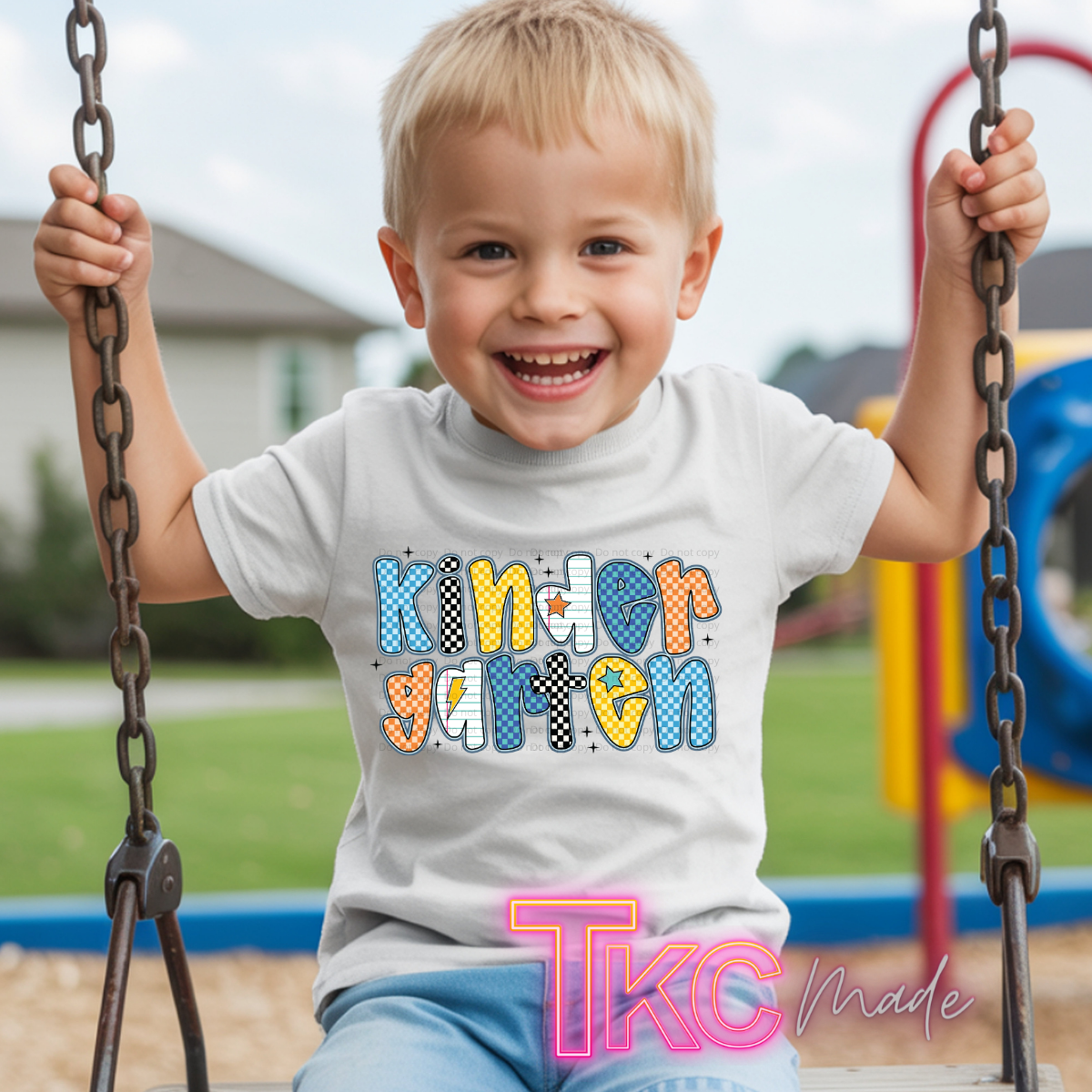 A young smiling boy sitting on a playground swing wearing a white t-shirt with a colorful 'Kindergarten' graphic. The graphic features patterned letters in blue, yellow, orange, and black-and-white checkered designs.