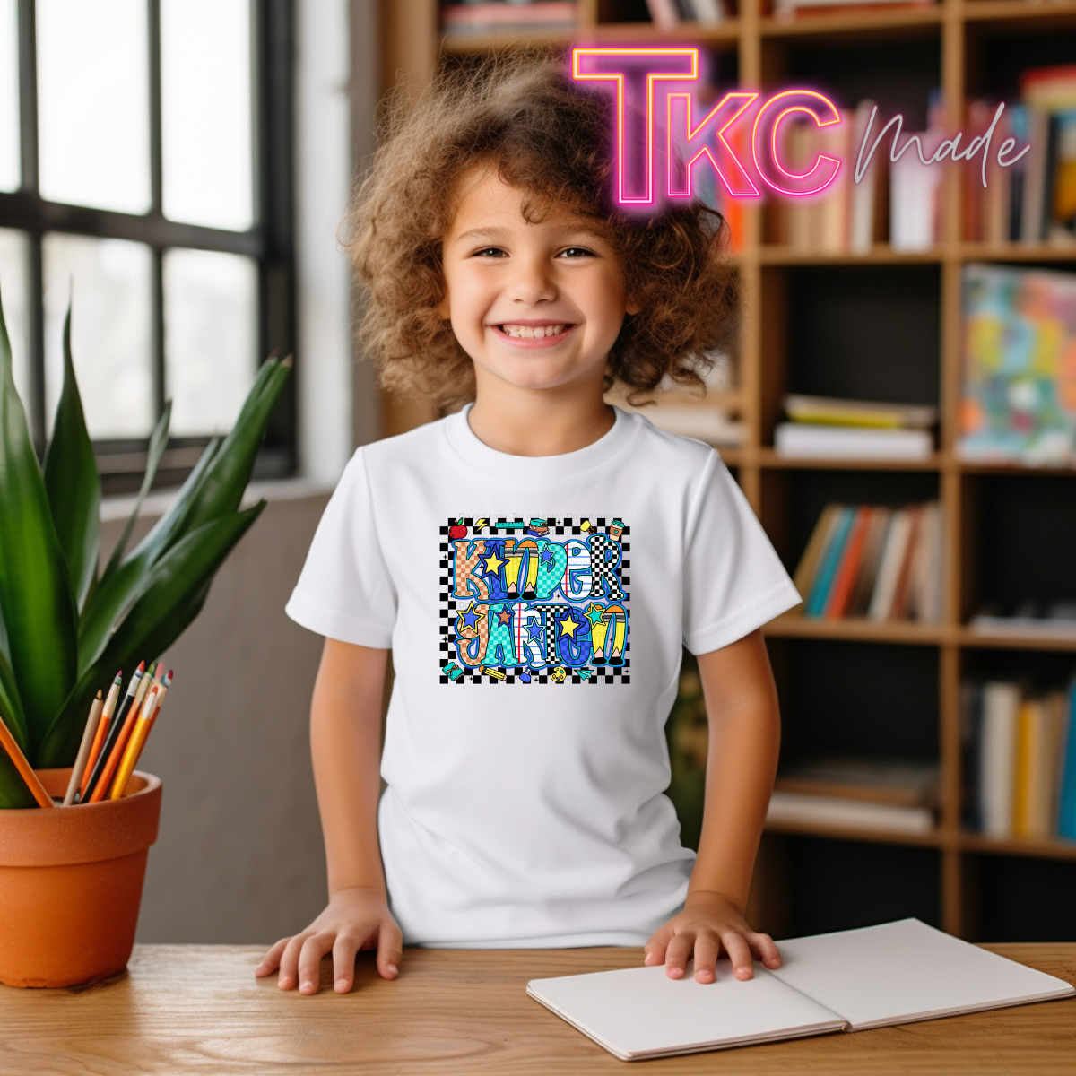 A smiling young girl with curly brown hair stands at a wooden desk wearing a white t-shirt with a colorful 'Kindergarten graphic. The background shows a cozy room with a bookshelf and a potted plant, and a pink 'TkC Made' watermark is visible in the top right corner.