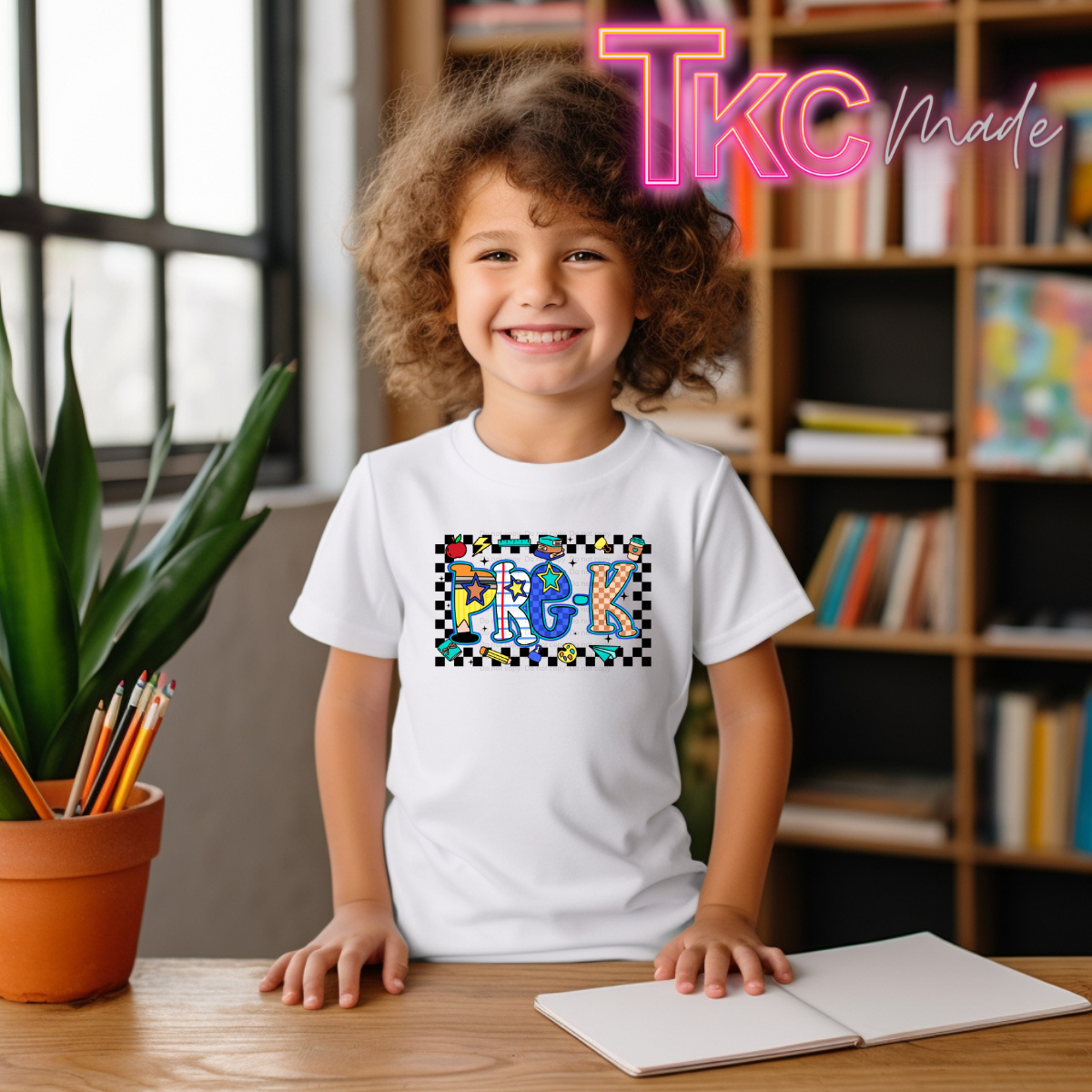 A smiling child with curly hair sits at a wooden desk wearing a white T-shirt with a colorful 'Pre-K graphic. The background shows a cozy room with a bookshelf and a potted plant, and a pink 'Tkc Made' watermark is visible in the top right corner.