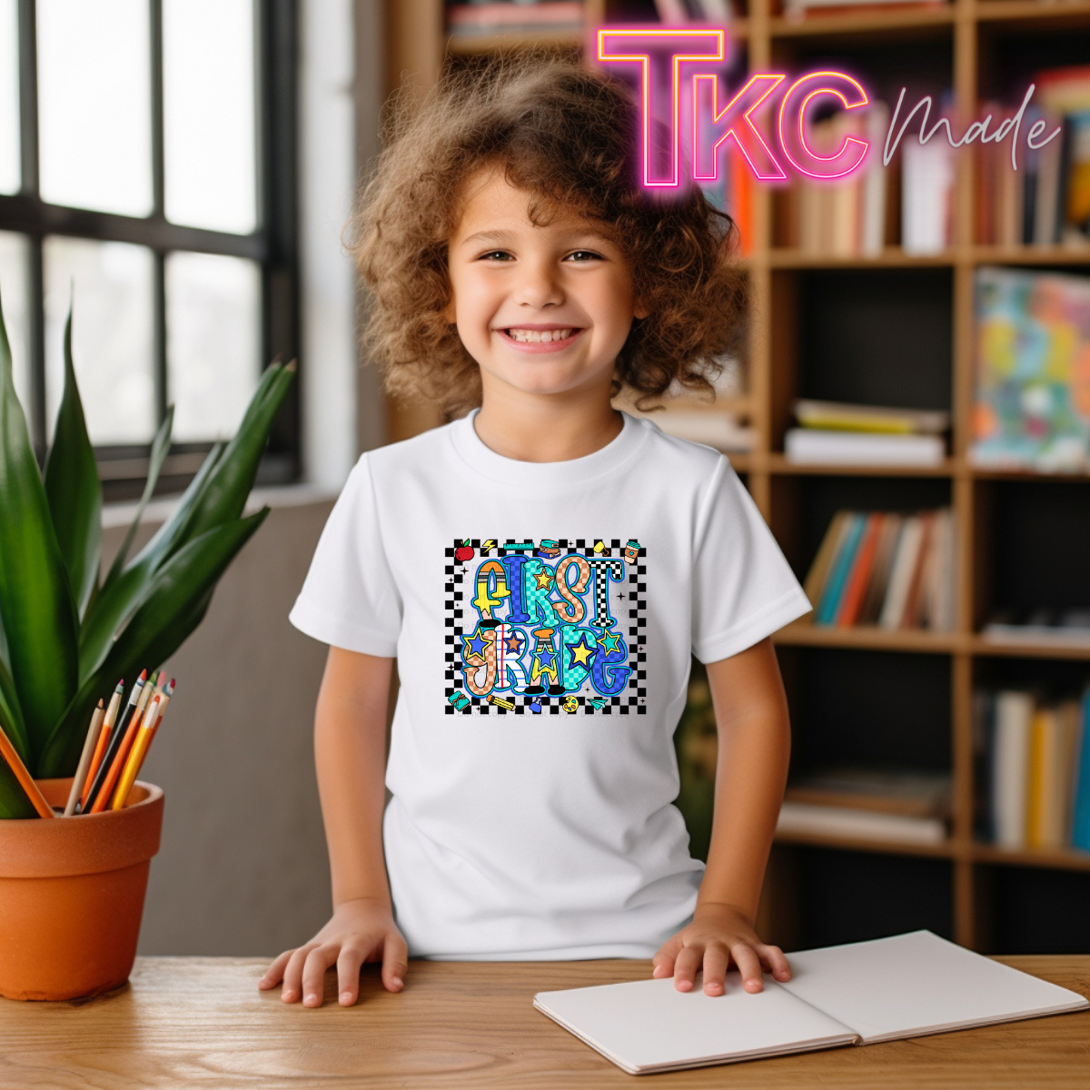 A smiling young child with curly hair sits at a wooden desk wearing a white T-shirt with a colorful 'First grade' graphic. The background shows a cozy indoor room with bookshelves, a potted plant, and a window.