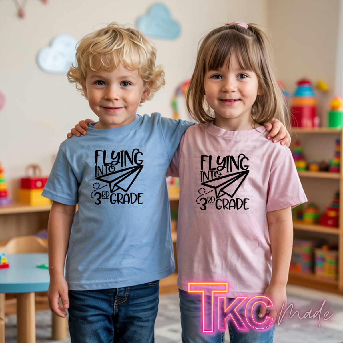 Two children standing together in a children's room, each wearing a graphic T-shirt. The boy wears a light blue shirt with black text reading 'FLYING INTO 1ST GRADE' and a paper airplane graphic, and the girl wears a light pink shirt with the same design.