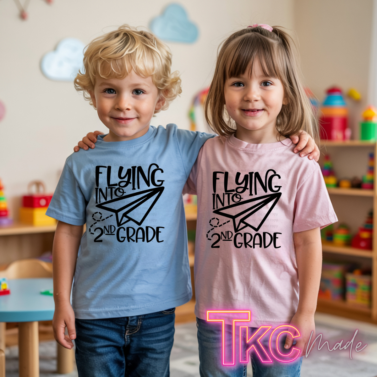 Two children standing in a playroom, each wearing a graphic T-shirt that reads 'Flying Into 2nd Grade' with a paper airplane design. The child on the left wears a light blue shirt, and the child on the right wears a light pink shirt.
