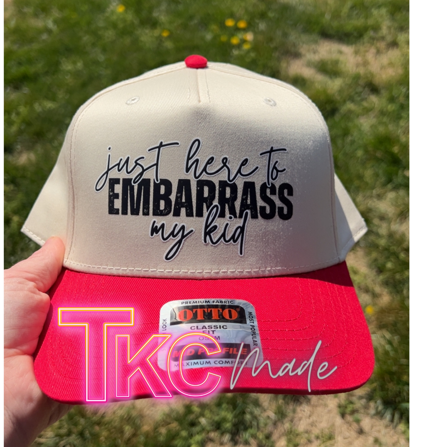 Cap with text 'just here to embarrass my kid' held against a grassy background
