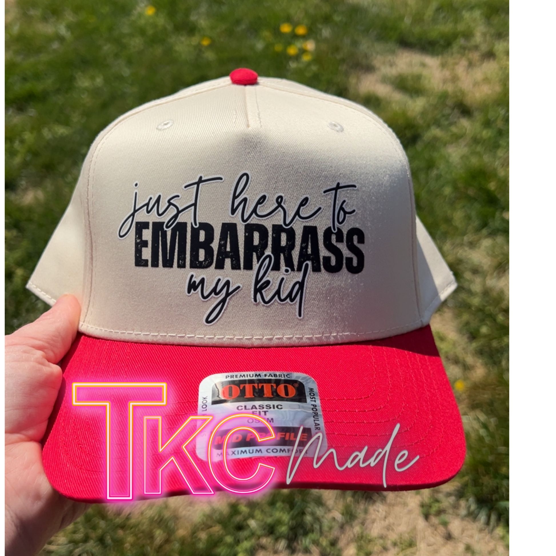 Cap with text 'just here to embarrass my kid' held against a grassy background