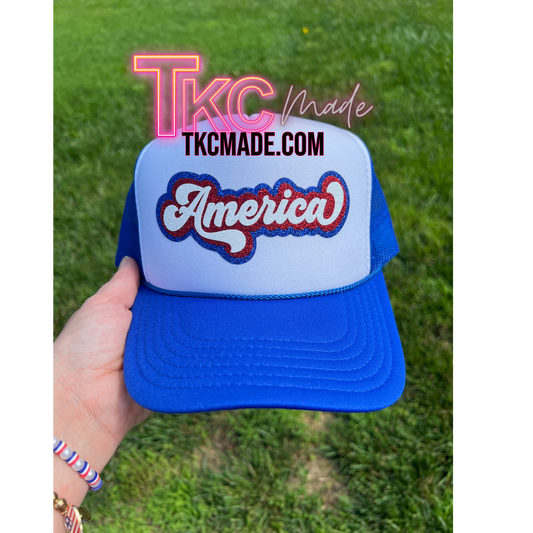 Blue and white cap with 'America' text held in front of a grassy background, featuring TKC Made branding.