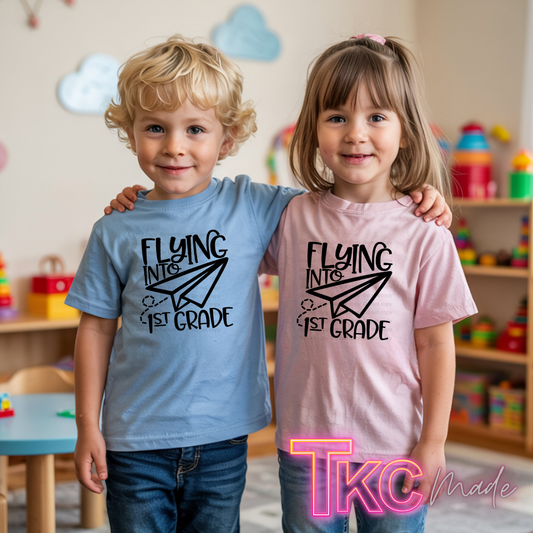 Two children standing together in a children's room, each wearing a graphic T-shirt. The boy wears a light blue shirt with black text reading 'FLYING INTO 1ST GRADE' and a paper airplane graphic, and the girl wears a light pink shirt with the same design.