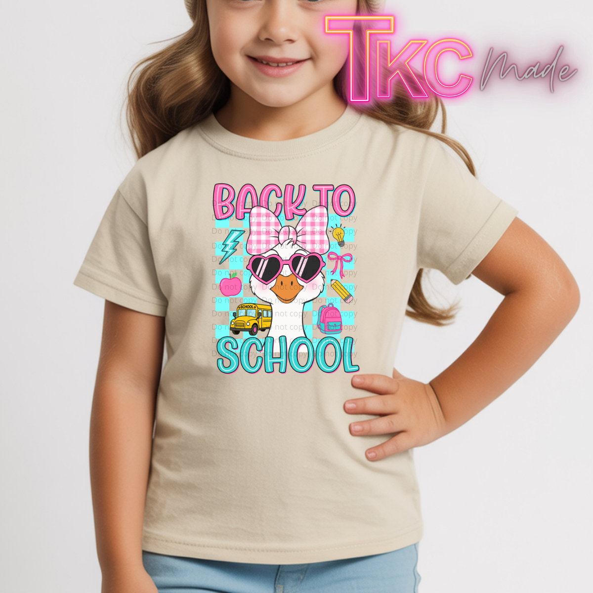 A young girl wearing a beige graphic t-shirt with a colorful 'Back to School' cartoon duck design. The shirt features a pink gingham bow, sunglasses, and school-themed icons.
