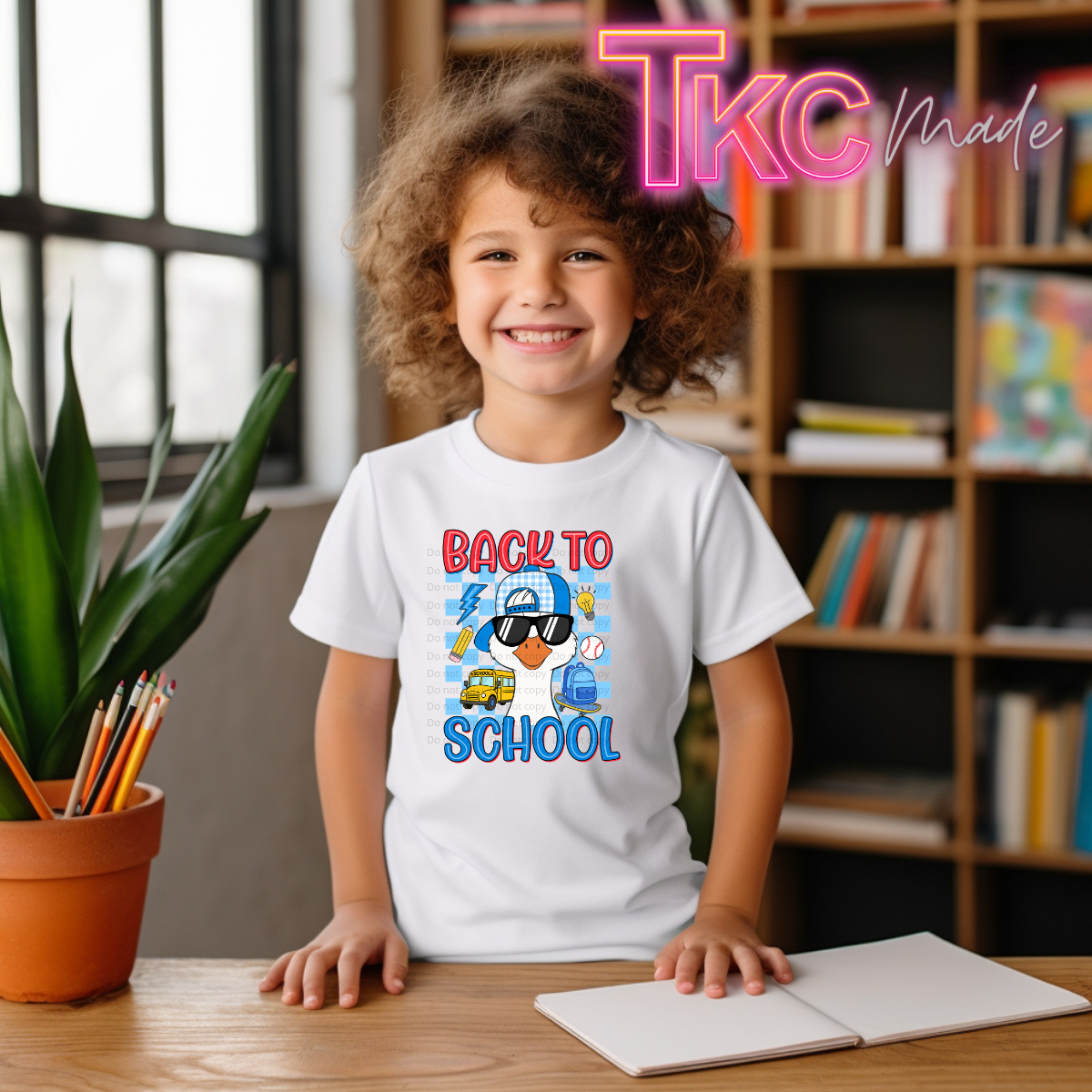 A smiling young girl with curly brown hair stands at a wooden desk wearing a white graphic T-shirt that reads 'BACK TO SCHOOL' with a cartoon illustration. The background shows a bookshelf, window, and potted plant, with a pink 'TKC Made' watermark overlay in the top right.