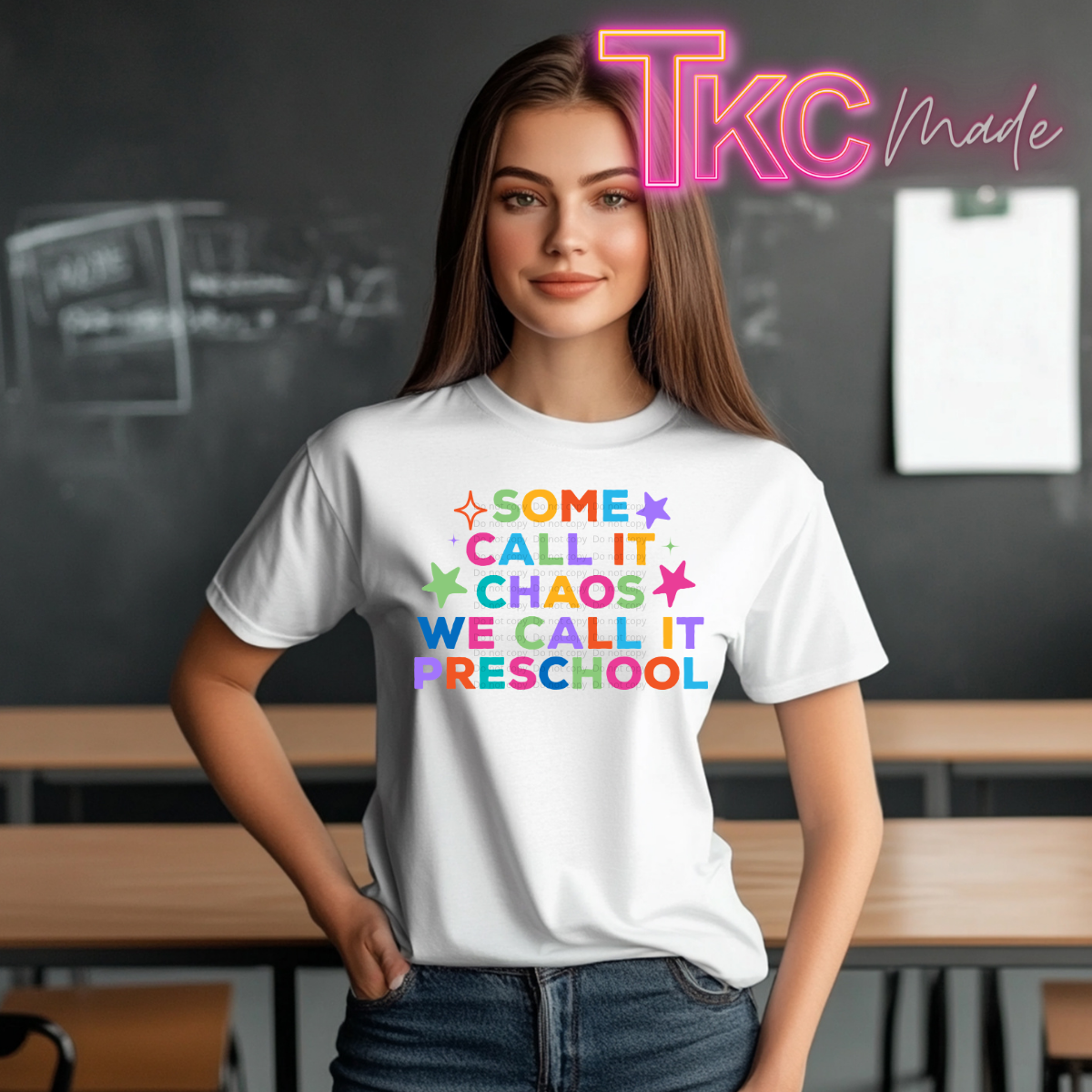 A woman wearing a white graphic t-shirt with the text 'SOME CALL IT CHAOS WE CALL IT PRESCHOOL' in colorful rainbow letters, standing in a classroom setting with a chalkboard background. A neon-style 'TKC made' logo is visible in the top right corner.