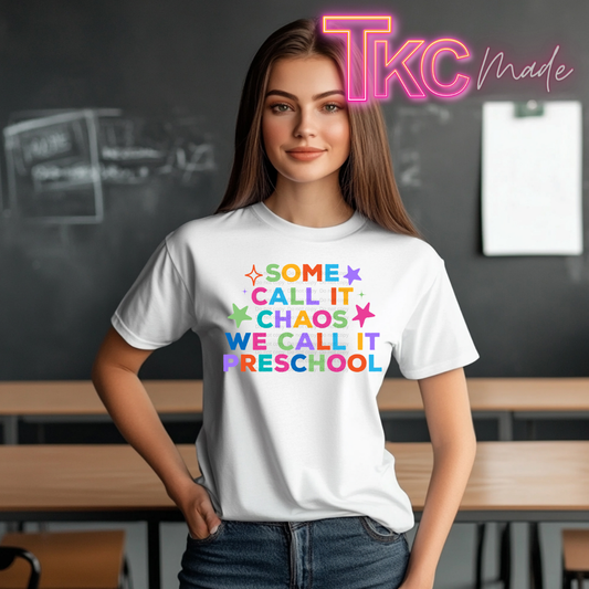 A woman wearing a white graphic t-shirt with the text 'SOME CALL IT CHAOS WE CALL IT PRESCHOOL' in colorful rainbow letters, standing in a classroom setting with a chalkboard background. A neon-style 'TKC made' logo is visible in the top right corner.