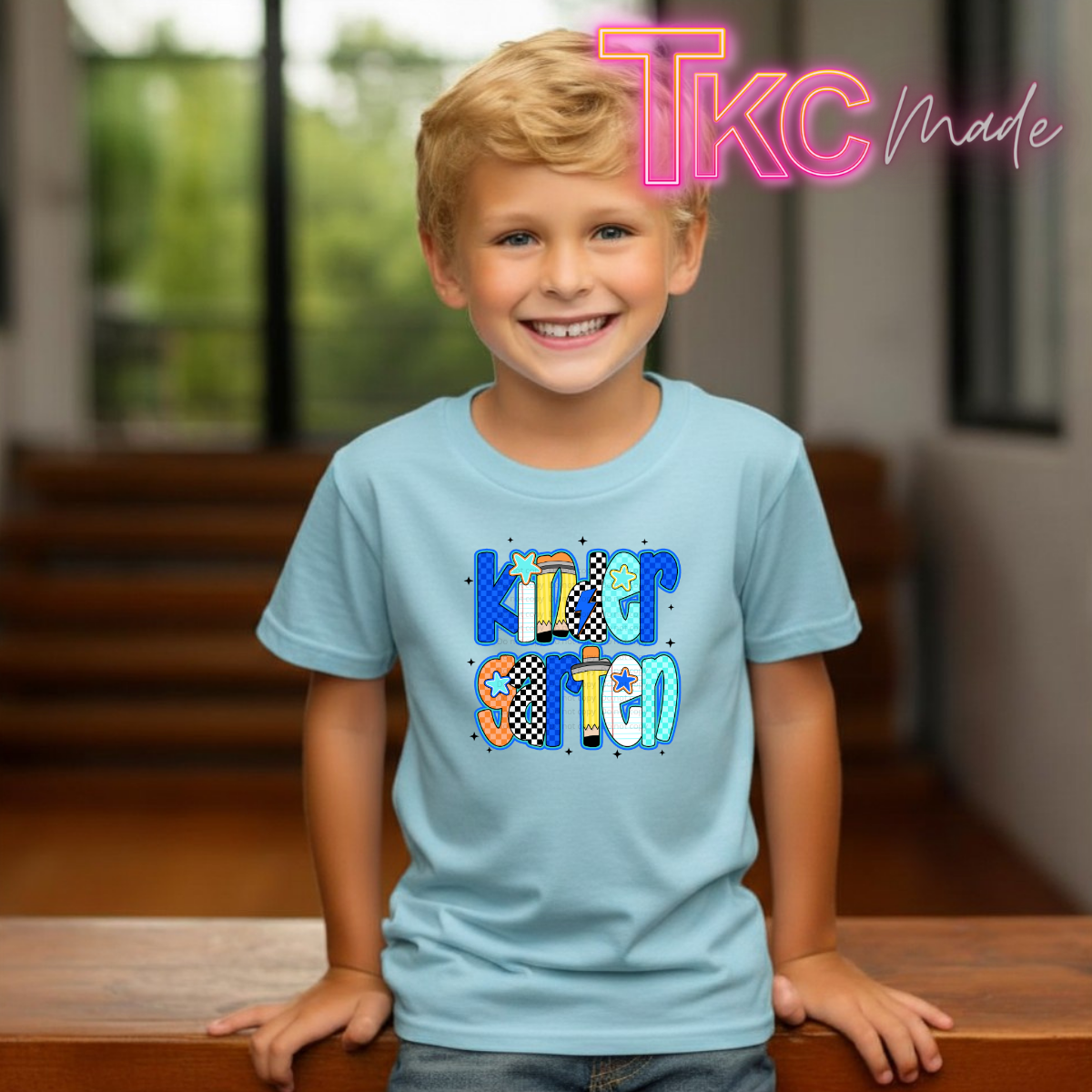 A young boy smiling and wearing a light blue t-shirt with a colorful 'Kindergarten' graphic on the front. The background shows a softly blurred indoor setting with wooden stairs and windows, and a pink 'TKC Made' watermark is visible in the top right corner.