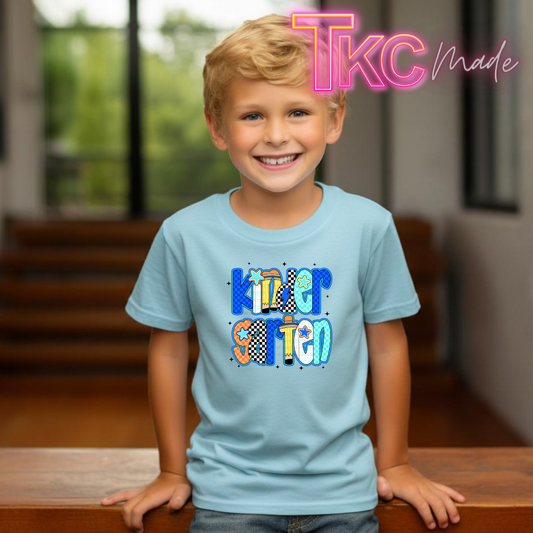 A young boy smiling and wearing a light blue t-shirt with a colorful 'Kindergarten' graphic on the front. The background shows a softly blurred indoor setting with wooden stairs and windows, and a pink 'TKC Made' watermark is visible in the top right corner.