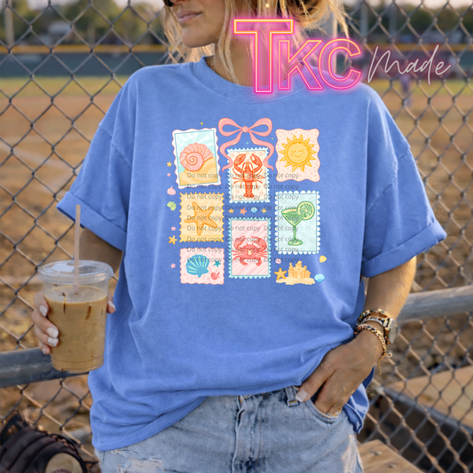 Person wearing a blue t-shirt with colorful beach graphics and holding a drink, standing in front of a chain-link fence.