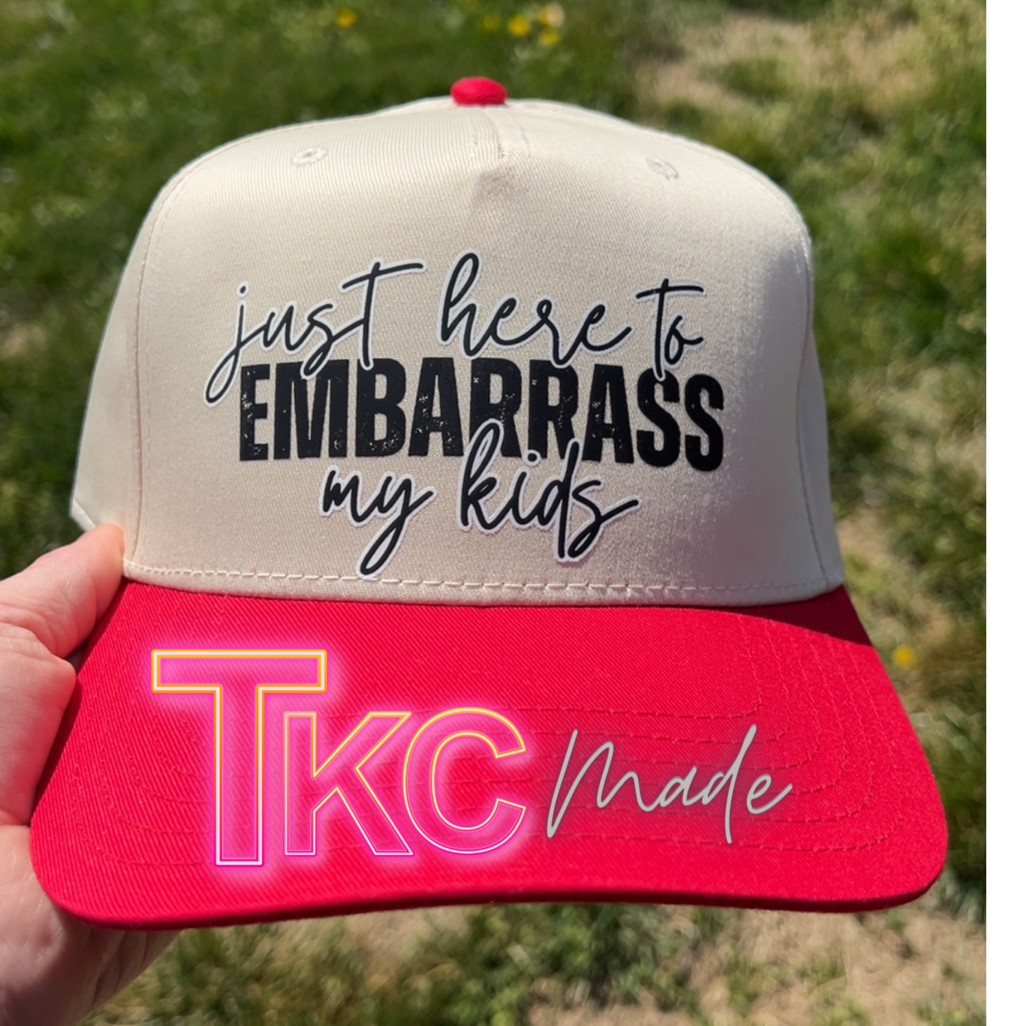 Cap with text 'just here to embarrass my kids' and 'TKC made' held in front of a grassy background