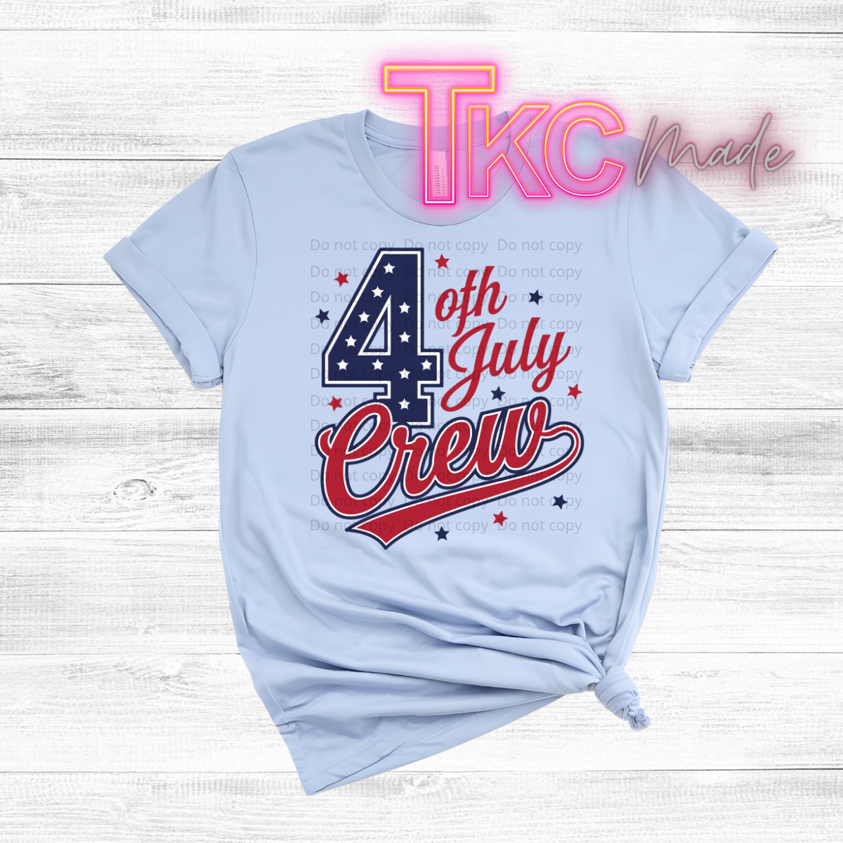Light blue t-shirt with '4th of July Crew' text on a wooden surface, featuring the brand 'TKC Made'.