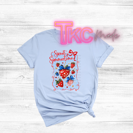 Light blue t-shirt with colorful strawberry design and 'TKC Made' branding on a wooden surface.