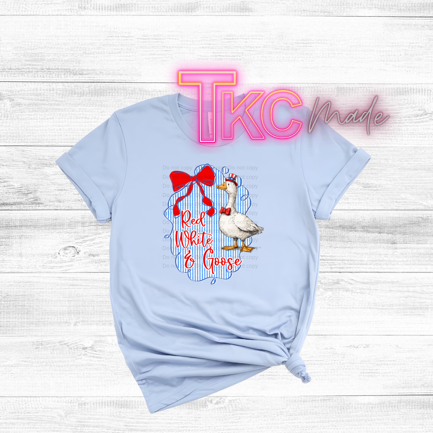 Light blue t-shirt with a goose graphic and text on a wooden surface, featuring 'TKC Made' branding.