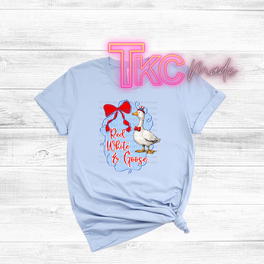 Light blue t-shirt with a goose graphic and text on a wooden surface, featuring 'TKC Made' branding.