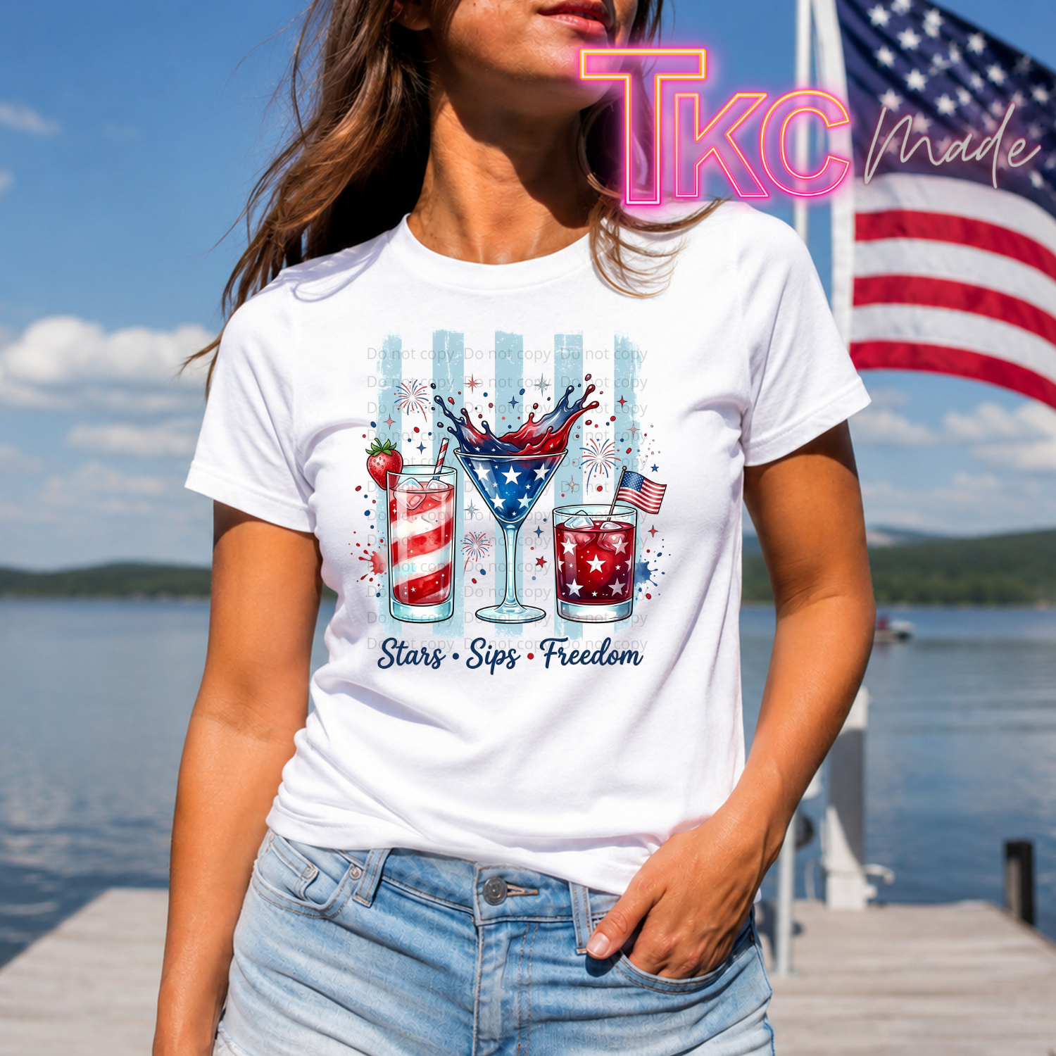 Person wearing a white t-shirt with a patriotic design  stars sips and freedom by a lake with an American flag in the background.