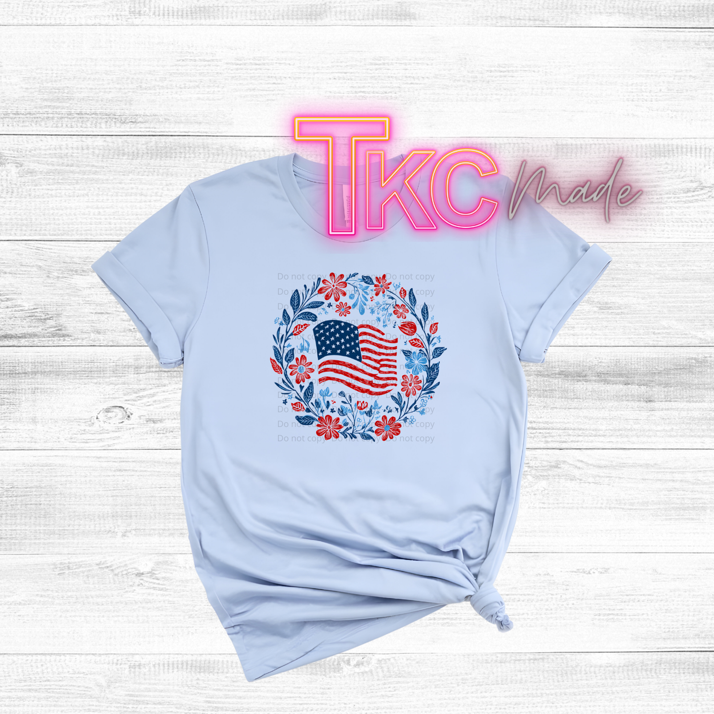 Light blue t-shirt with American flag design on a wooden surface, featuring 'TKC Made' branding.