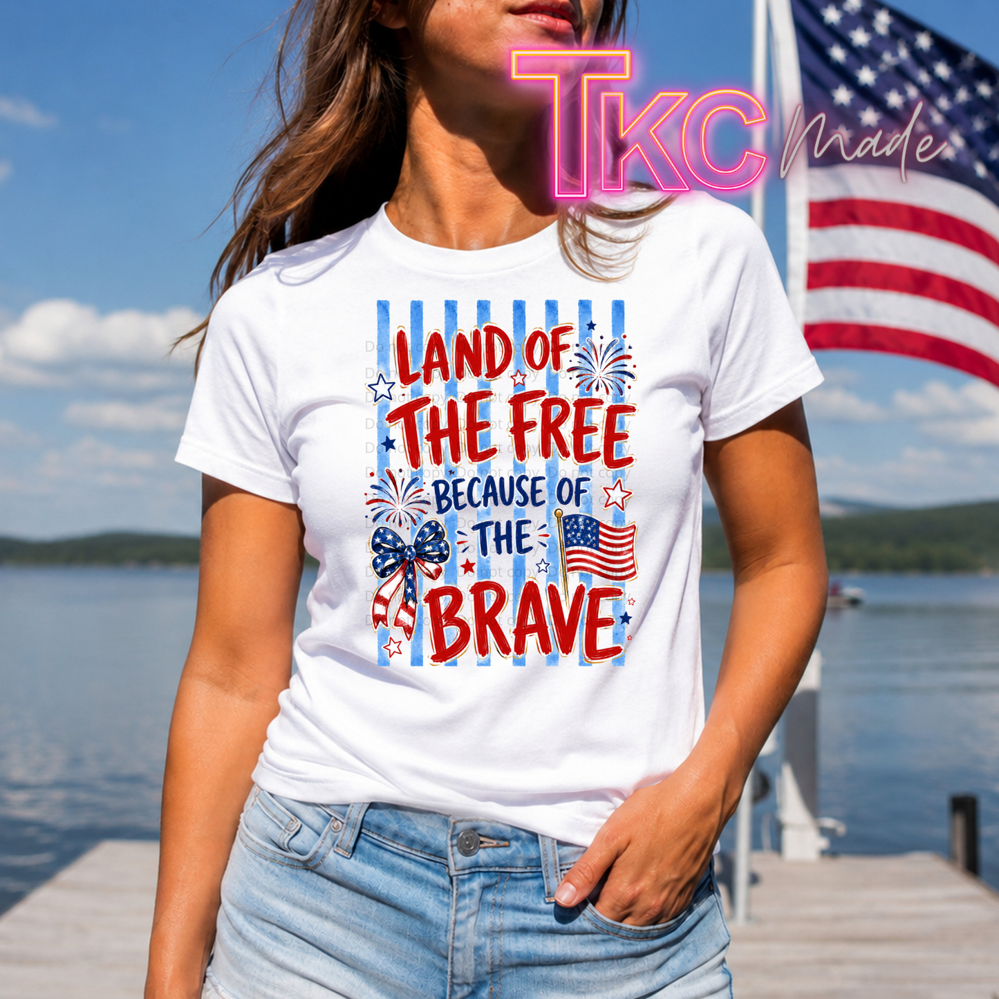 Person wearing a white t-shirt with a patriotic design  land of the free because of the brave by a lake with an American flag in the background.
