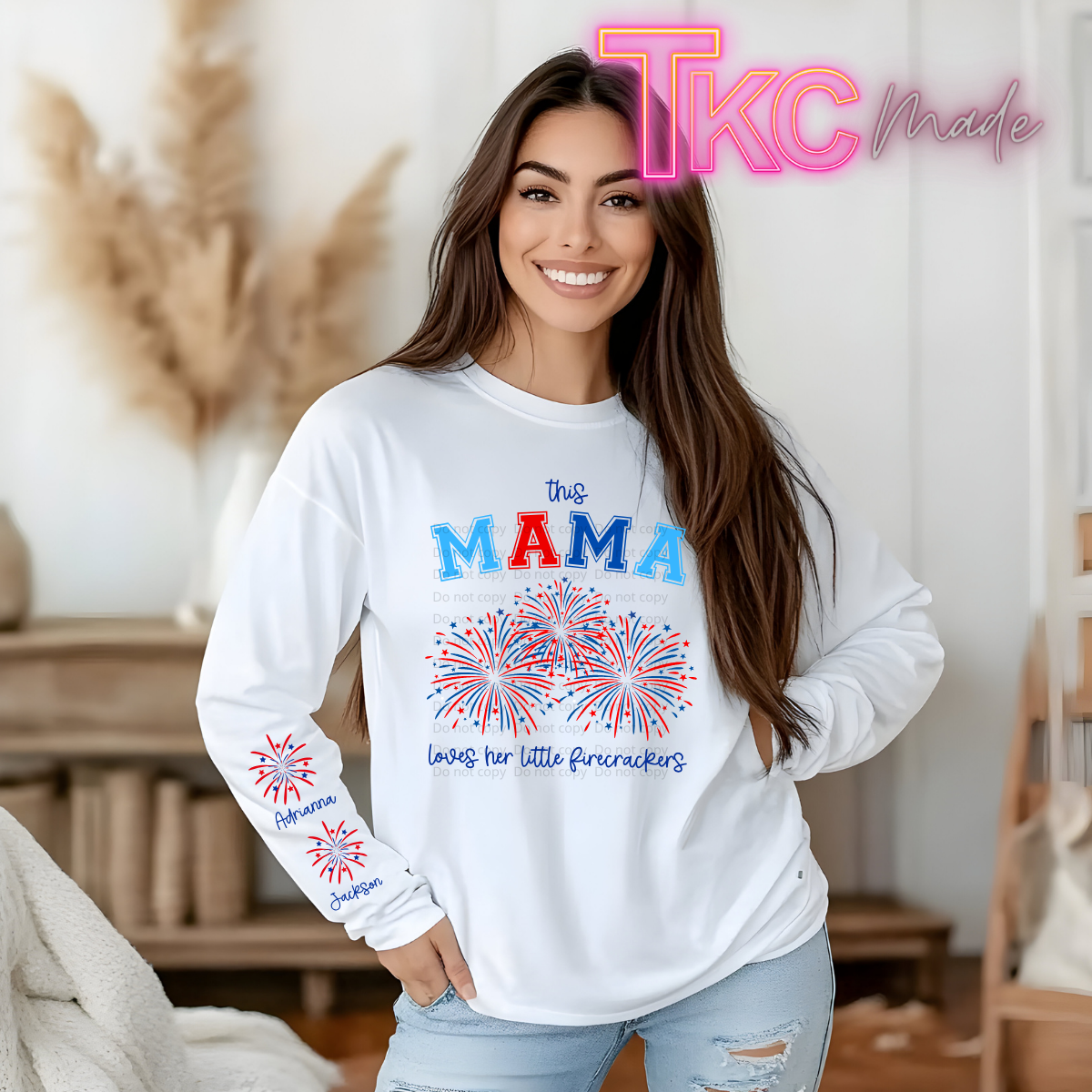 Woman wearing a white sweatshirt with 'this MAMA loves her little firecrackers' text and fireworks design, indoors.