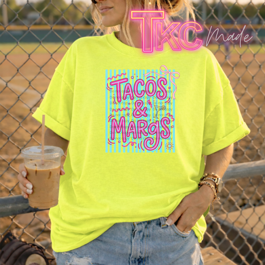 Person wearing a bright yellow t-shirt with 'Tacos & Marg' text, holding a drink, standing in front of a chain-link fence.