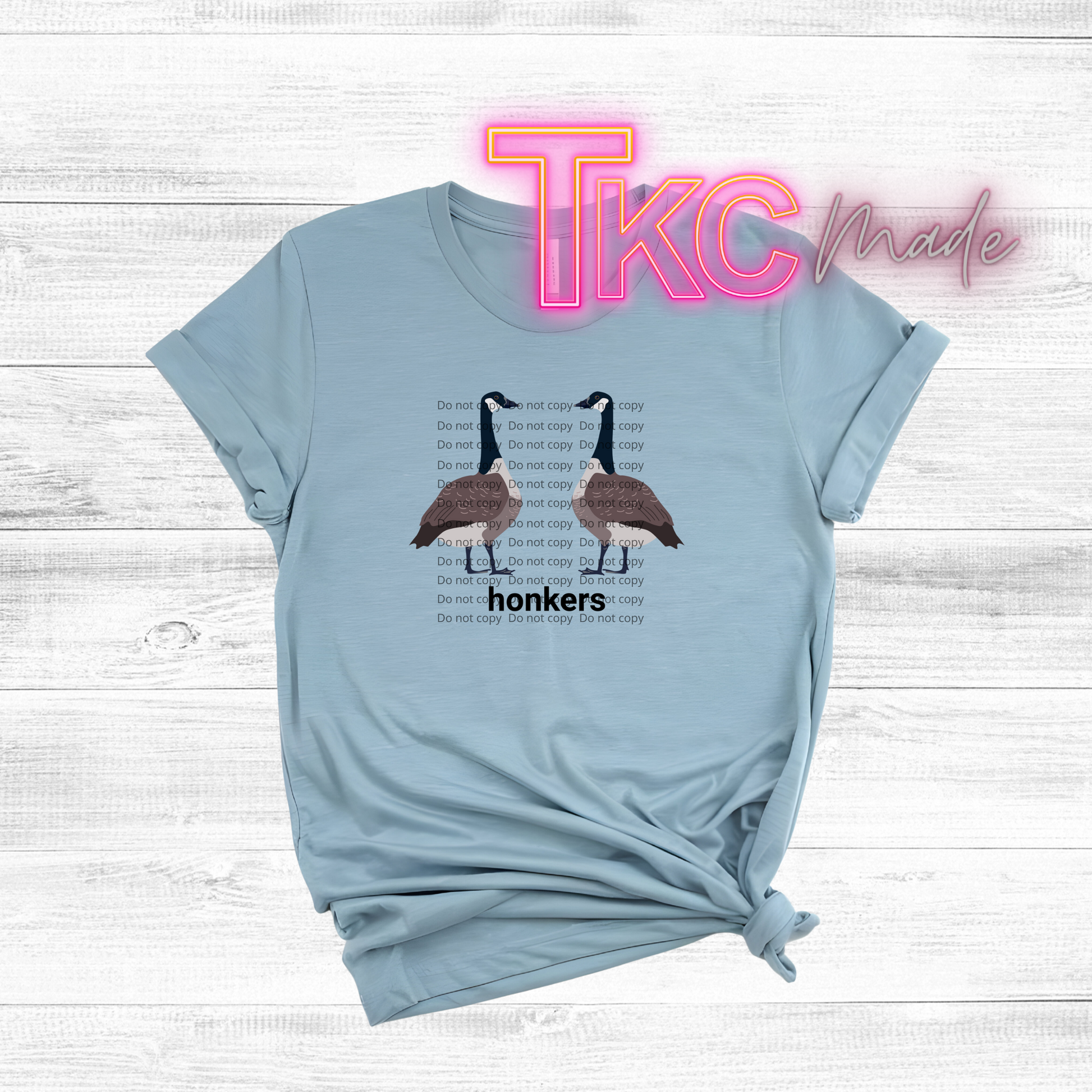 Light blue t-shirt with goose graphic and text on a wooden surface, featuring 'TKC Made' branding.