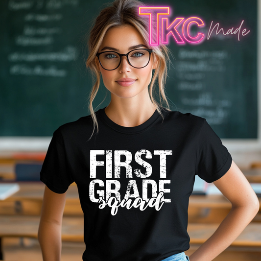A woman wearing a black graphic t-shirt with the text 'FIRST GRADE squad' in white, standing in a classroom setting with a chalkboard in the background. A 'TKC made' watermark is visible in the top right corner.