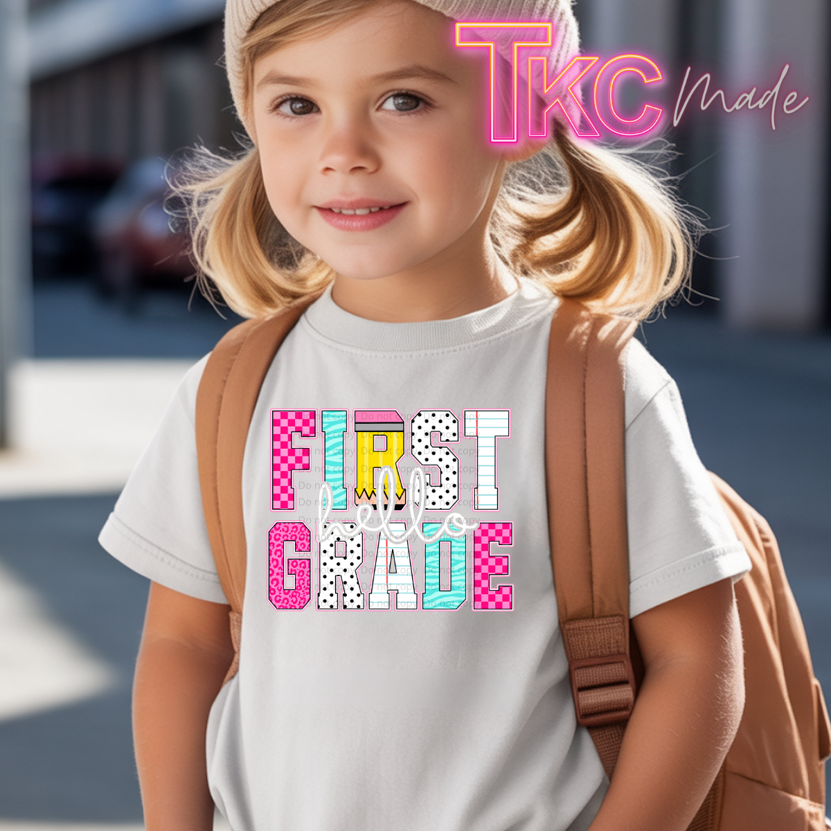 A young girl with blonde pigtails and a tan backpack wearing a white t-shirt with a colorful 'Hello FIRST Grade' graphic, set against a blurred outdoor street background with a 'TkC Made' watermark.