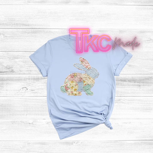 Light blue t-shirt with a floral bunny design on a wooden surface, featuring 'TKC Made' text.