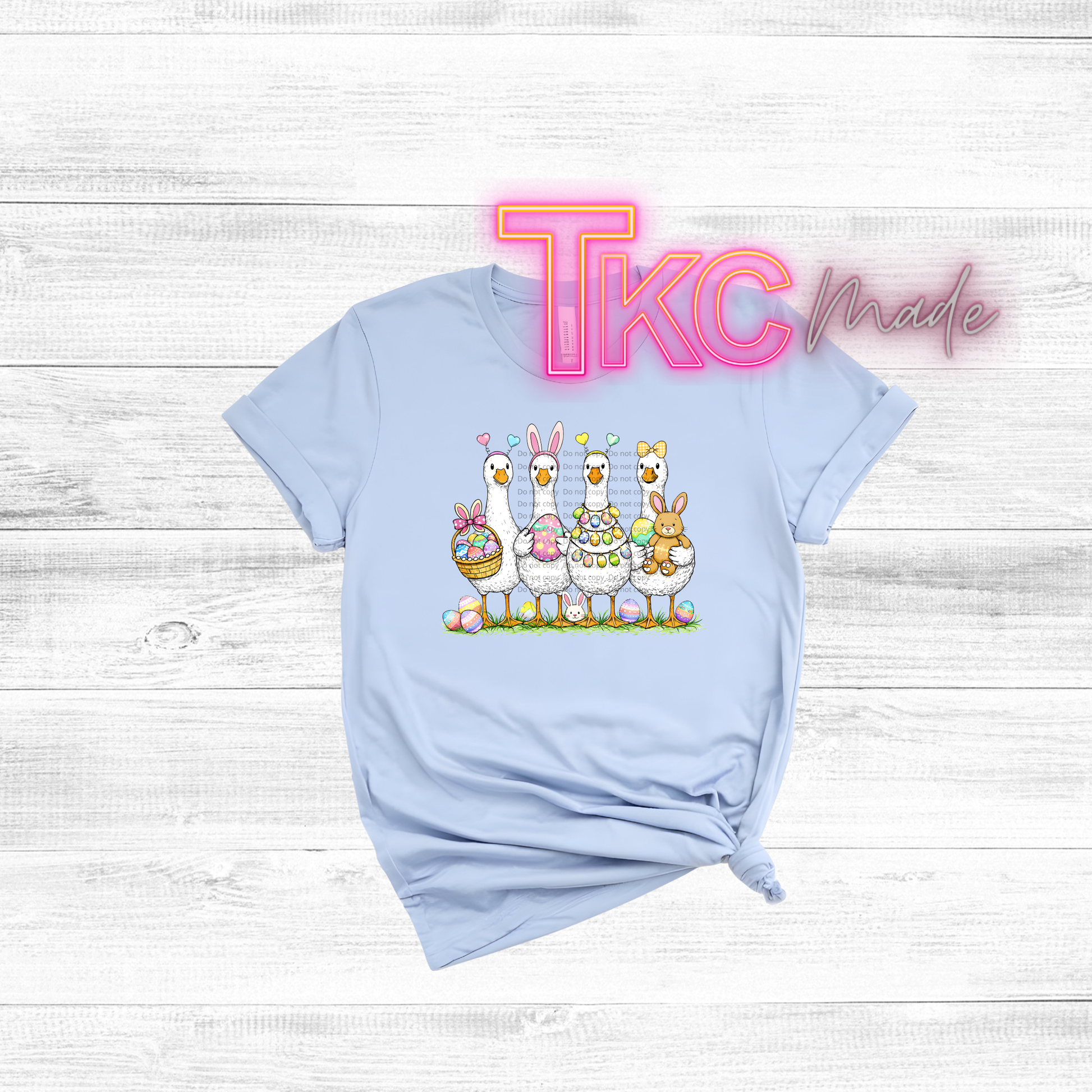 Light blue t-shirt with cartoon goose and Easter eggs on a white wooden background, featuring 'TKC Made' text.