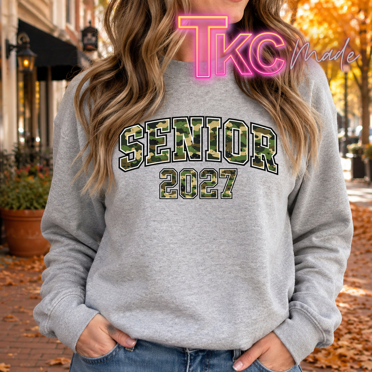 A woman wearing a heather gray crewneck sweatshirt with 'SENIOR 2027' printed in a green camouflage pattern on the front, set against an outdoor autumn street background.