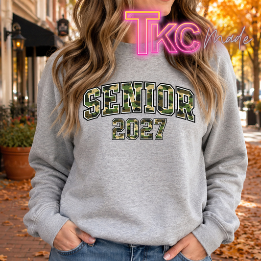 A woman wearing a heather gray crewneck sweatshirt with 'SENIOR 2027' printed in a green camouflage pattern on the front, set against an outdoor autumn street background.