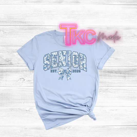 Light blue t-shirt with 'Senior 2026' design on a wooden surface, featuring 'TKC Made' text.