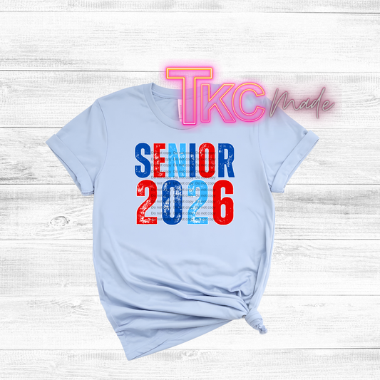 Light blue t-shirt with 'Senior 2026' text in red and blue on a white wooden background.