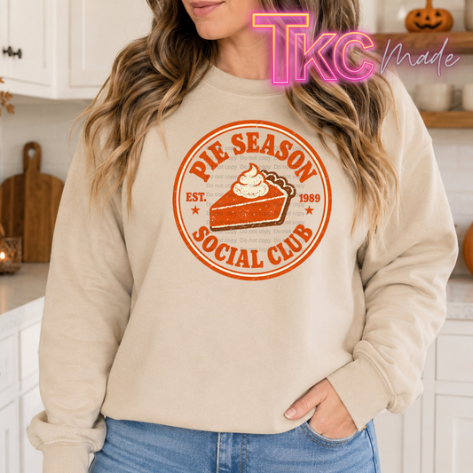 A woman wearing a beige crewneck sweatshirt with a retro-style orange graphic that reads 'PIE SEASON SOCIAL CLUB' around an illustration of a slice of pumpkin pie. The photo is taken in a bright kitchen setting, with a 'TKC Made' watermark overlay in the top right corner.