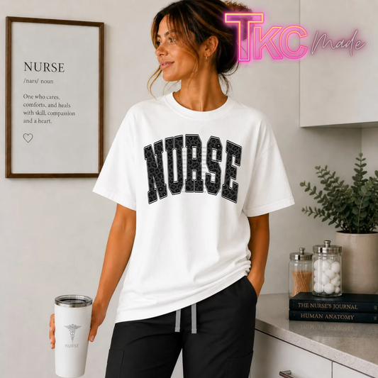 A woman wearing a white crewneck t-shirt with 'NURSE' printed in black arched letters across the chest, holding a white travel tumbler. She is standing in a kitchen setting with a framed print on the wall, a countertop with books and jars, and a plant.