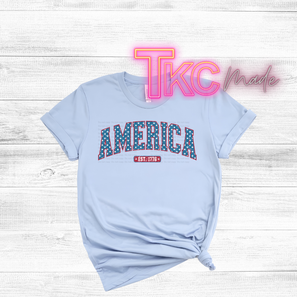 Light blue t-shirt with 'AMERICA EST. 1776' text on a wooden surface, featuring 'TKC made' branding.