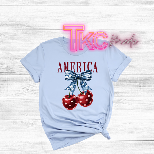 Light blue t-shirt with 'AMERICA' and cherry design on a wooden surface, featuring 'TKC Made' branding.